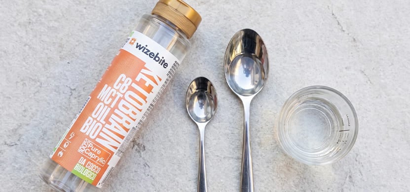 Flacone di olio MCT C8 Ketobrain Wizebite da cocco biologico affiancato da due cucchiai in acciaio e un bicchierino dosatore.